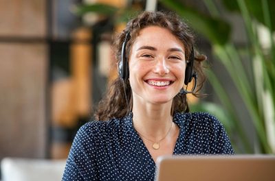 Portrait of smiling casual businesswoman working in a creative office. Smiling friendly woman working as call center agent for online support. Young woman in video call with headphones looking at camera.